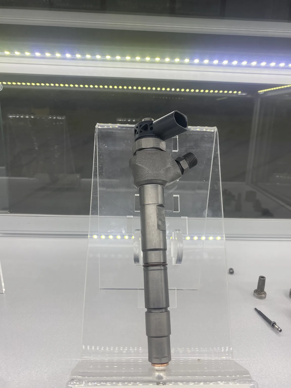 Gray diesel injector display sample on an acrylic stand inside a glass cabinet.